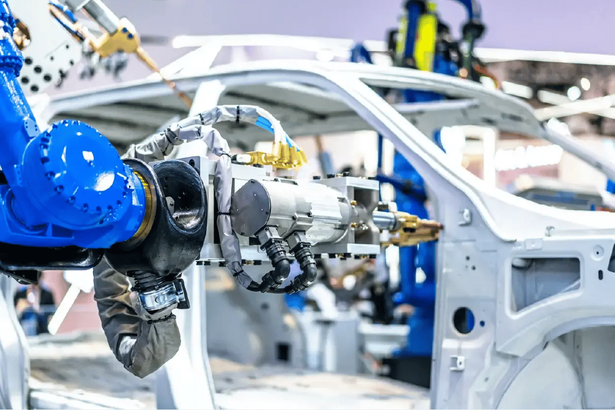 Robotic arm welding a car body part outdoors, with workers in safety gear in the background.