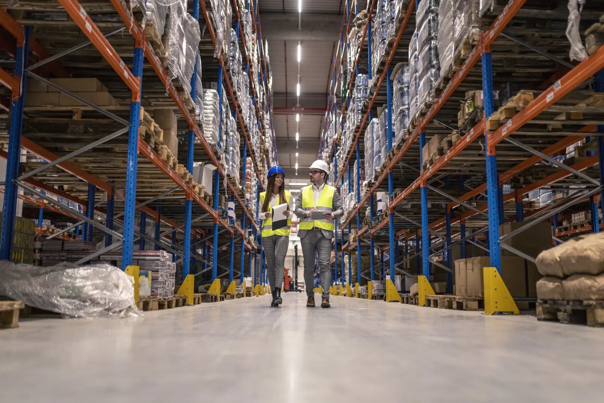 workers-with-hardhats-reflective-jackets-walking-through-big-warehouse-aisle-checking-goods-condition