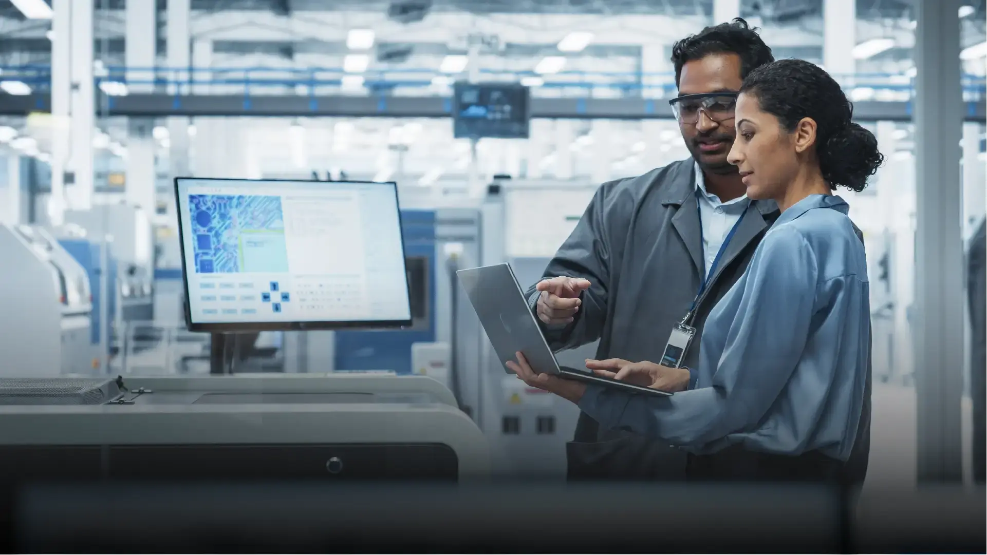 Two professionals discussing technical information on a laptop in a modern industrial facility, with machines and monitors in the background, representing equipment integration.